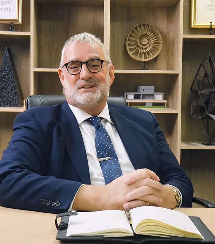An investment advisor smiling behind a desk, with his notebook over the table and bhind him, a cabinet filled with sculptures, diplomas and a 3d house mockuo